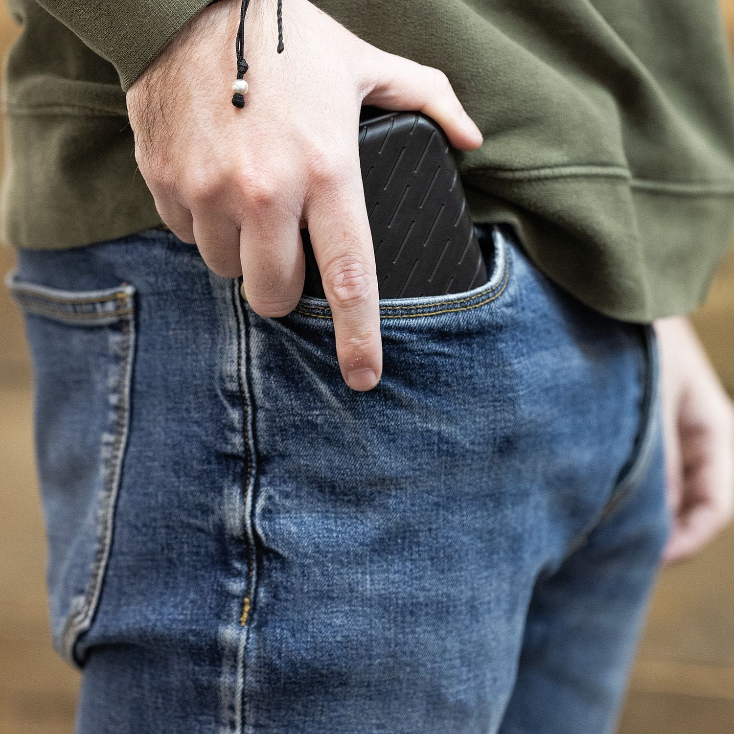 Étui à fléchettes Harrows Slimcase Eine Person steckt ein schwarzes Etui in die Hosentasche. Sie trägt eine grüne Jacke und eine blaue Jeans.