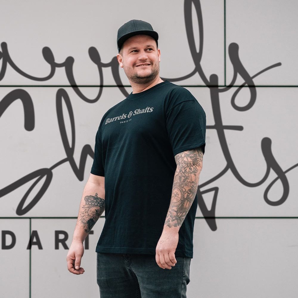 T-Shirt Barils et Arbres - Noir Ein Mann trägt ein schwarzes T-Shirt mit der Aufschrift „Barrels & Shafts“. Er steht vor einer Wand mit passender Schrift im Hintergrund.