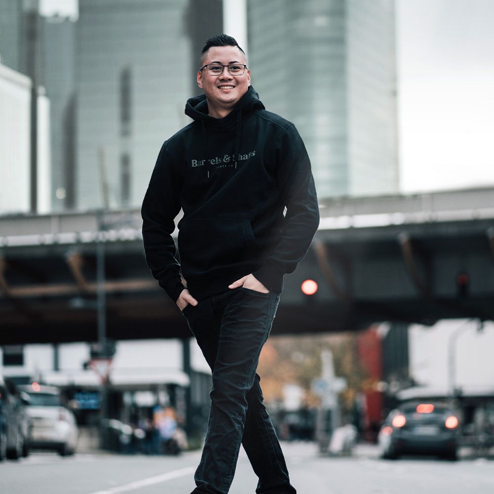 Sweat à capuche Barrels and Shafts - Noir Eine Person trägt einen schwarzen "Barrels and Shafts" Hoodie und steht entspannt auf einer Straße. Im Hintergrund sind unscharfe Gebäude und Autos zu sehen.