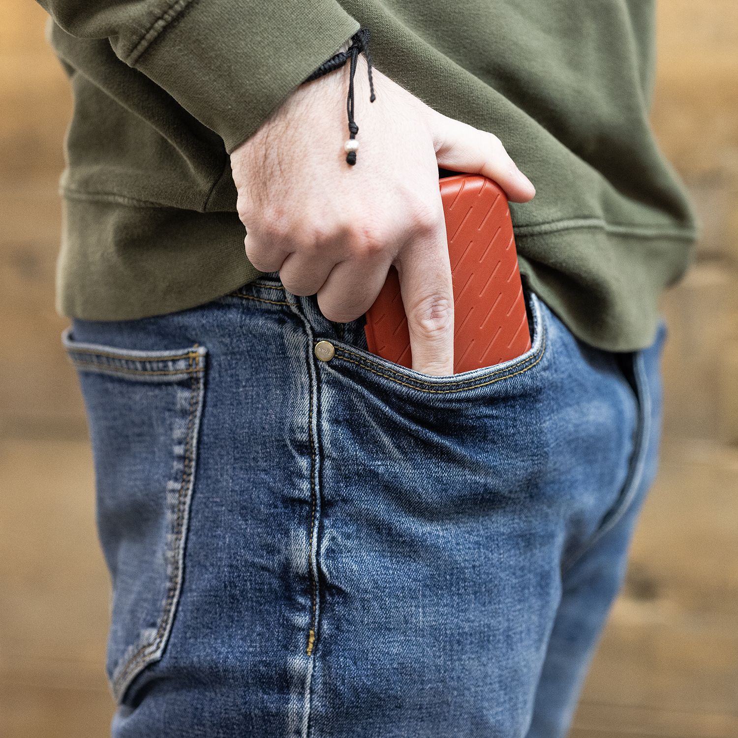 Eine Person steckt ein rotes Etui in die Hosentasche. Sie trägt eine blaue Jeans und einen grünen Pullover.