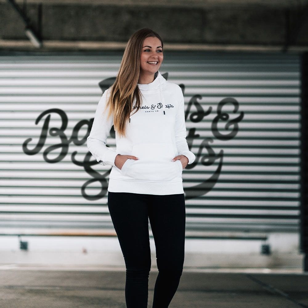 Sweat à capuche Femme Barrels and Shafts - Blanc Eine Frau trägt einen weißen "Barrels and Shafts" Damen Hoodie. Sie steht vor einem Hintergrund mit großem Schriftzug und lächelt.