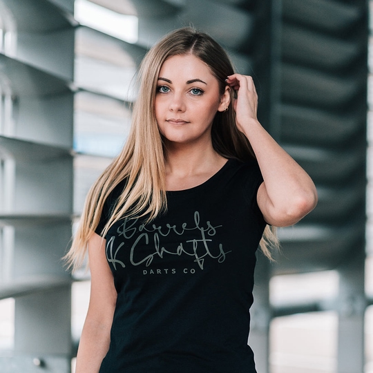 T-Shirt Femme Barrels and Shafts - Noir Eine Frau trägt ein schwarzes T-Shirt mit der Aufschrift „Barrels and Shafts Darts Co“. Sie steht vor einem modernen, lichtdurchfluteten Hintergrund.