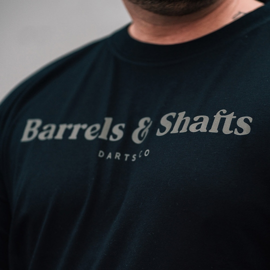 T-Shirt Barils et Arbres - Noir Das Bild zeigt ein schwarzes T-Shirt mit der Aufschrift "Barrels & Shafts". Darunter steht in kleineren Buchstaben "DARTSCO".