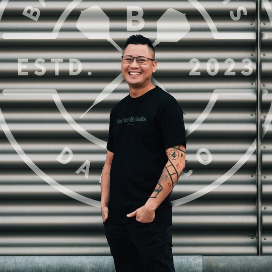 T-Shirt Barils et Arbres - Noir Ein Mann trägt ein schwarzes "Barrels and Shafts"-T-Shirt und steht vor einer metallenen Wand mit großem Logo. Er lächelt und hat markante Tattoos am rechten Arm.
