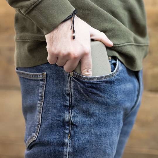 Étui à fléchettes Harrows Slimcase Eine Person steckt eine graue Darttasche in ihre Hosentasche. Die Person trägt eine blaue Jeans und einen olivgrünen Pullover.